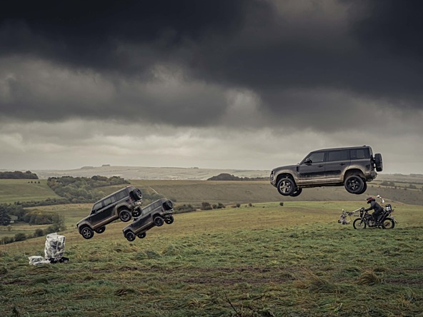 Land Rover soaring through the air while racing a motorcycle in a grassy field with dark clouds in the background